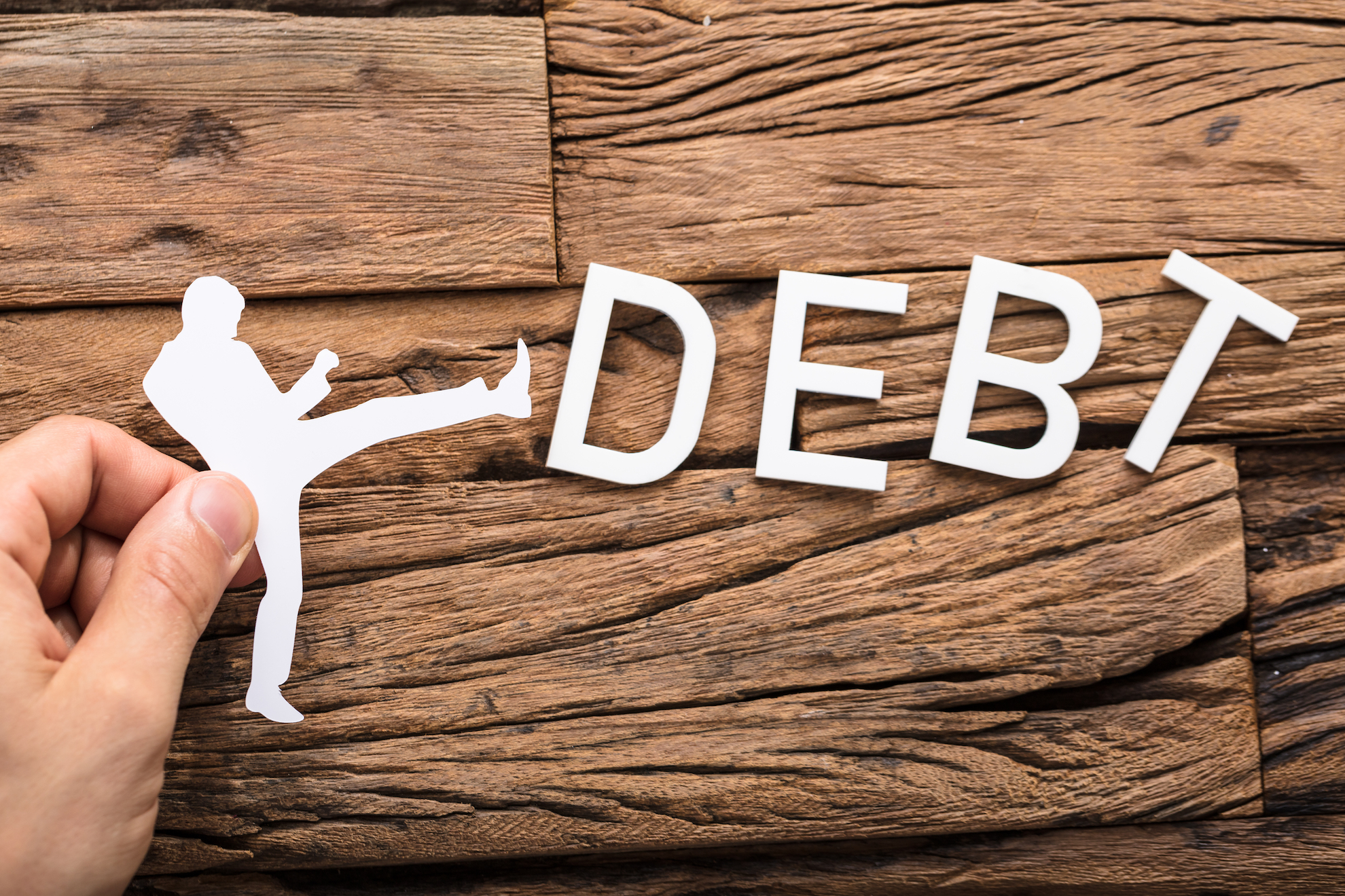 Hand Holding A Paper Cut Out Figure Kicking The Debt Word On Wooden Table
